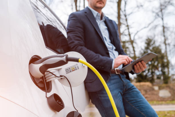 Business man standing near charging electric car or EV car and using tablet in the street at the sunlight.
